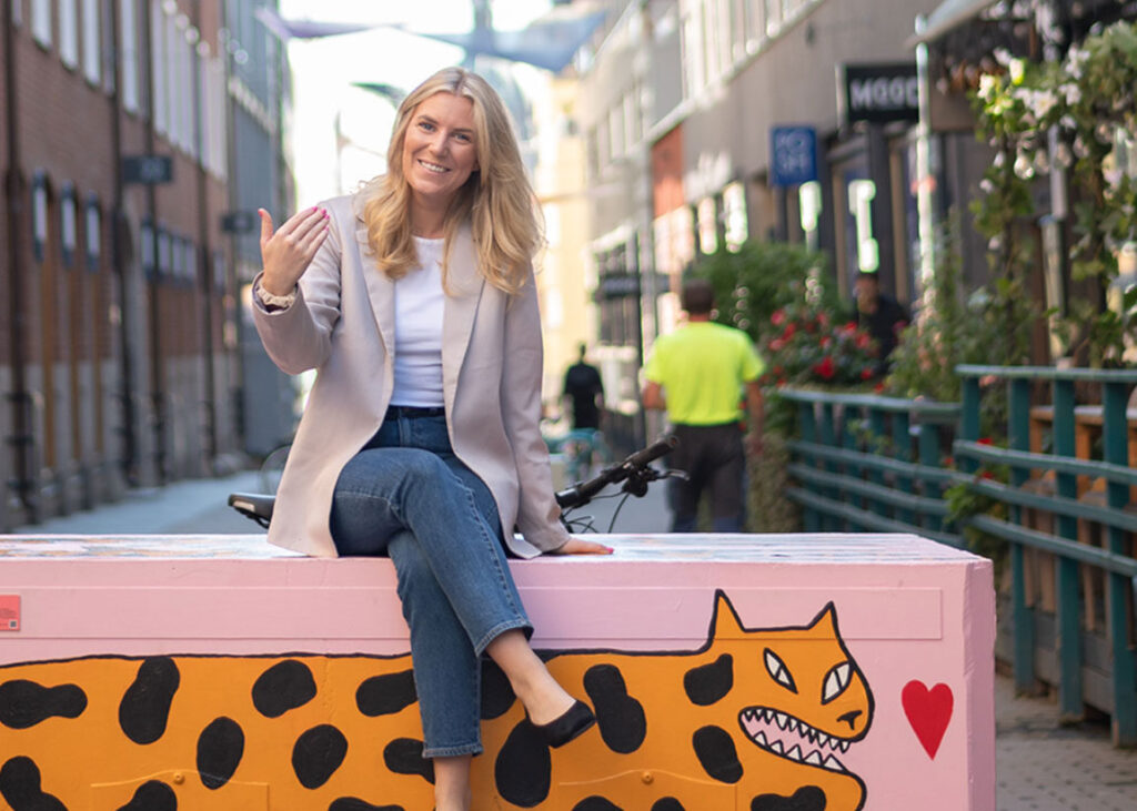 Amanda, rekryterare på Imprint Recruitment i Stockholm, sitter leende på en färgglad bänk med leopardmönster och ett hjärta, omgiven av en livlig stadsmiljö. Bilden förmedlar ett välkomnande och professionellt tillvägagångssätt inom rekrytering.